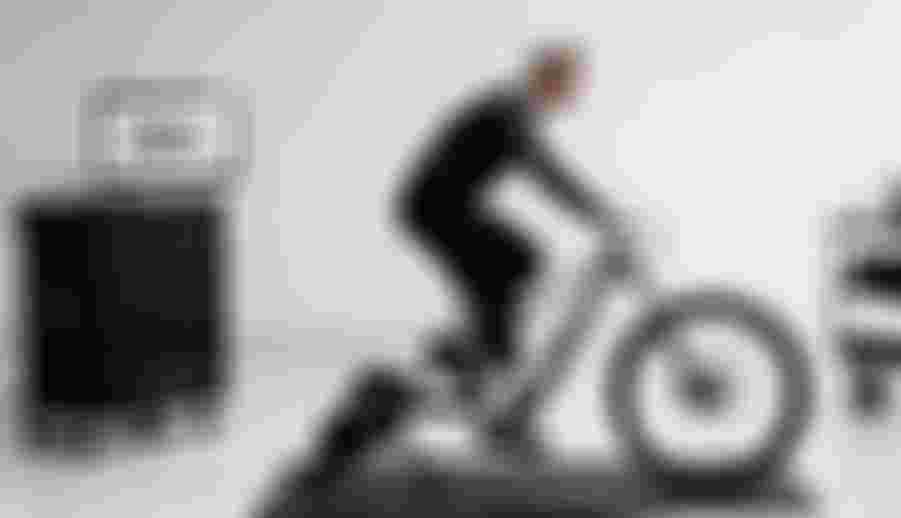 Male cyclist riding a mountain bike on an indoor trainer in a lab setting, with an SQlab saddle displayed on a monitor in the background.