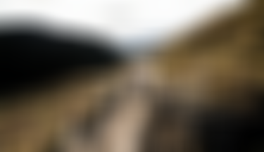 Two mountain bikers descending a flowing alpine singletrack trail, kicking up dust while cornering through a berm with wide mountain landscape in the background.