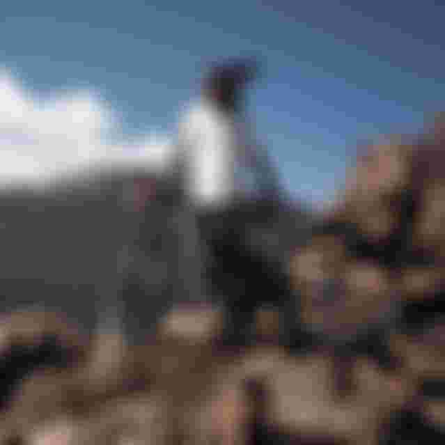 Mountain biker in gear stands with a bike on rocky terrain, overlooking a rugged landscape with a distant smoking volcano under a clear blue sky.