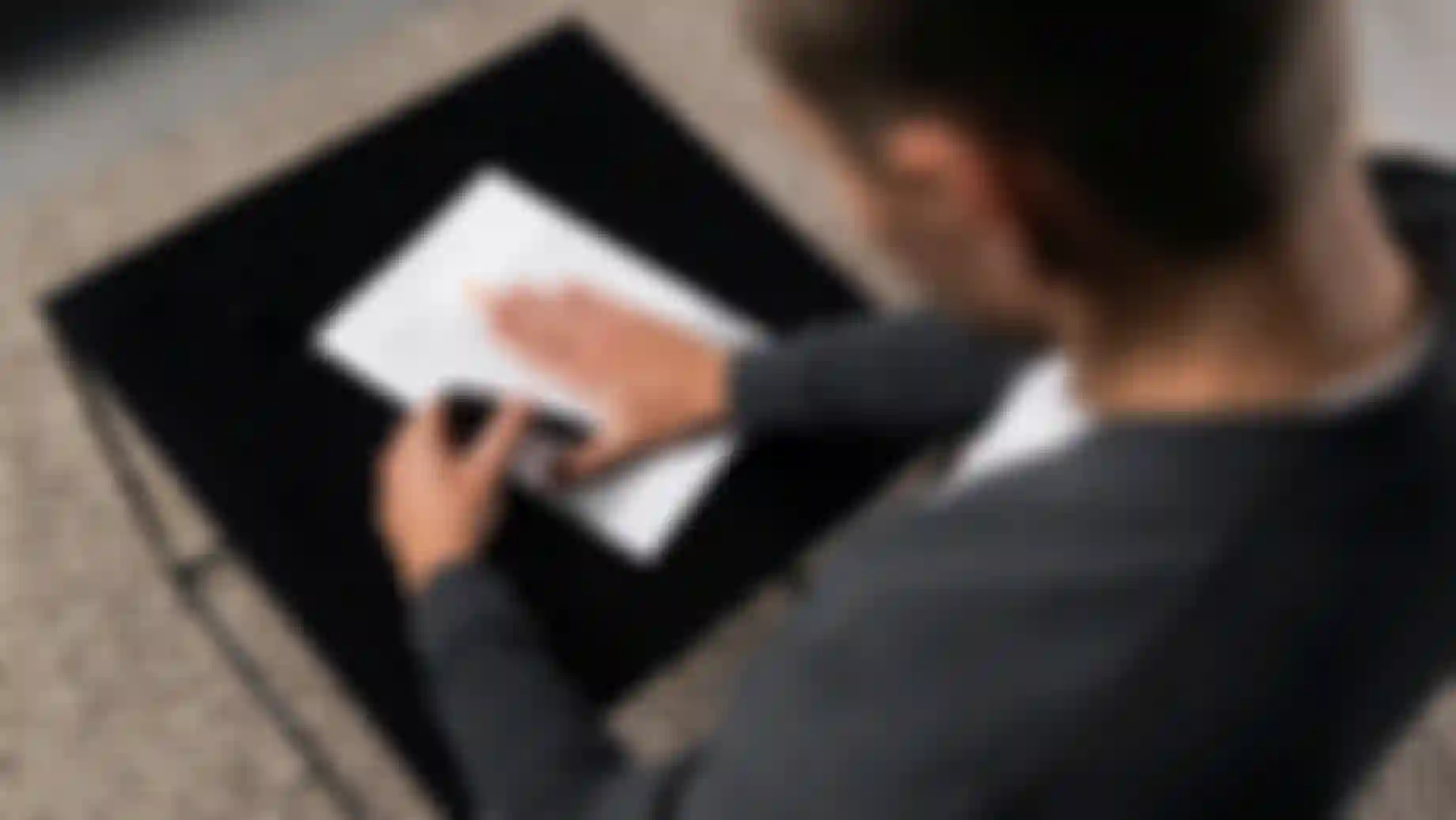 A person in a grey sweater uses a pen to mark a typed document on a black table, with a beige textured carpet underneath.