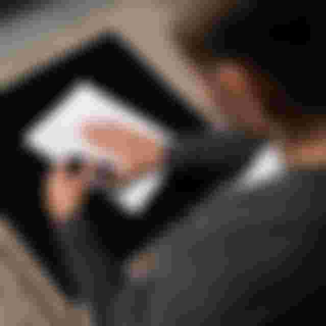 A person in a grey sweater uses a pen to mark a typed document on a black table, with a beige textured carpet underneath.