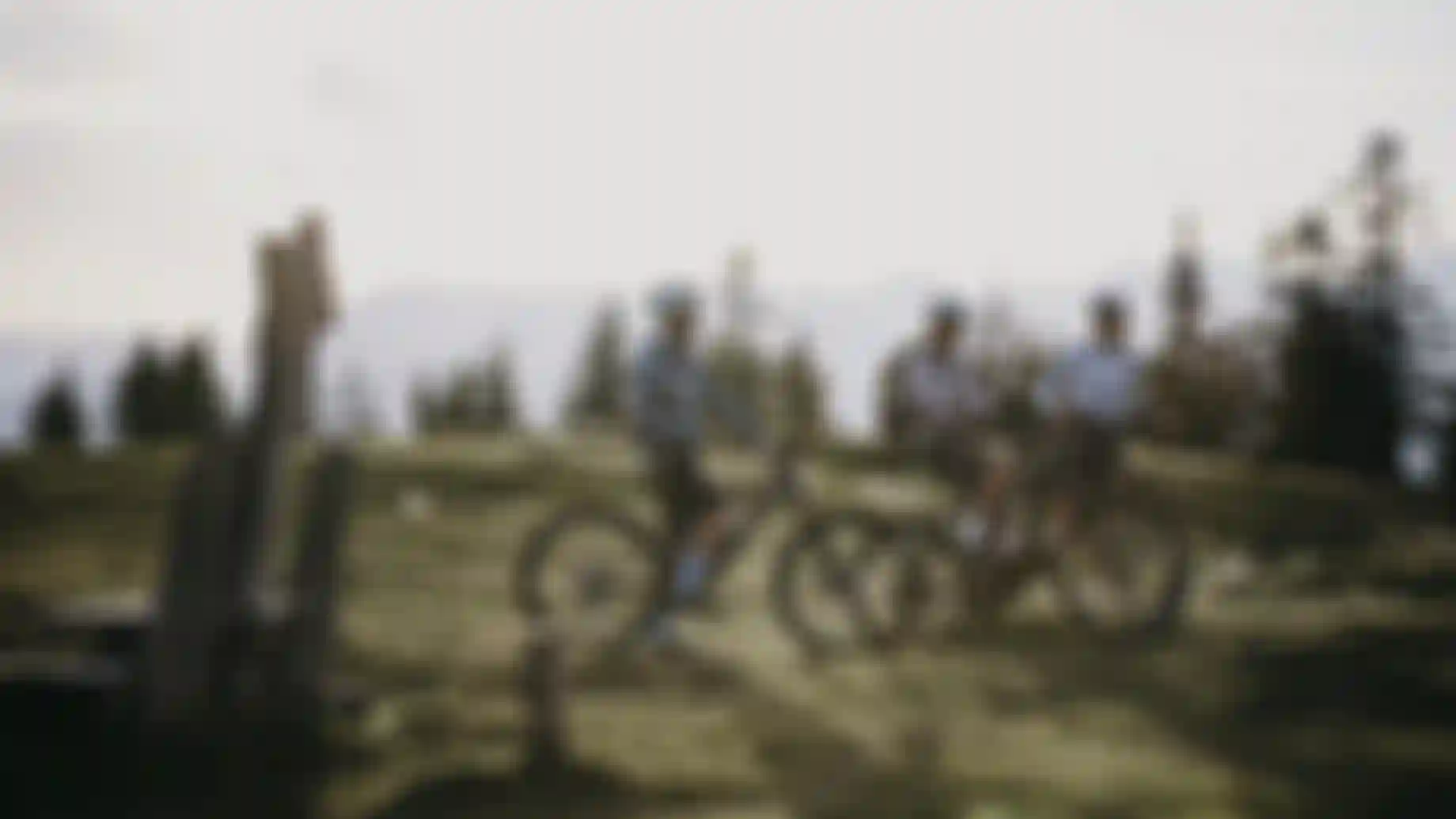 Three mountain bikers wearing helmets pause with their bikes on an alpine meadow at sunset, surrounded by trees and distant mountain silhouettes in soft golden light.