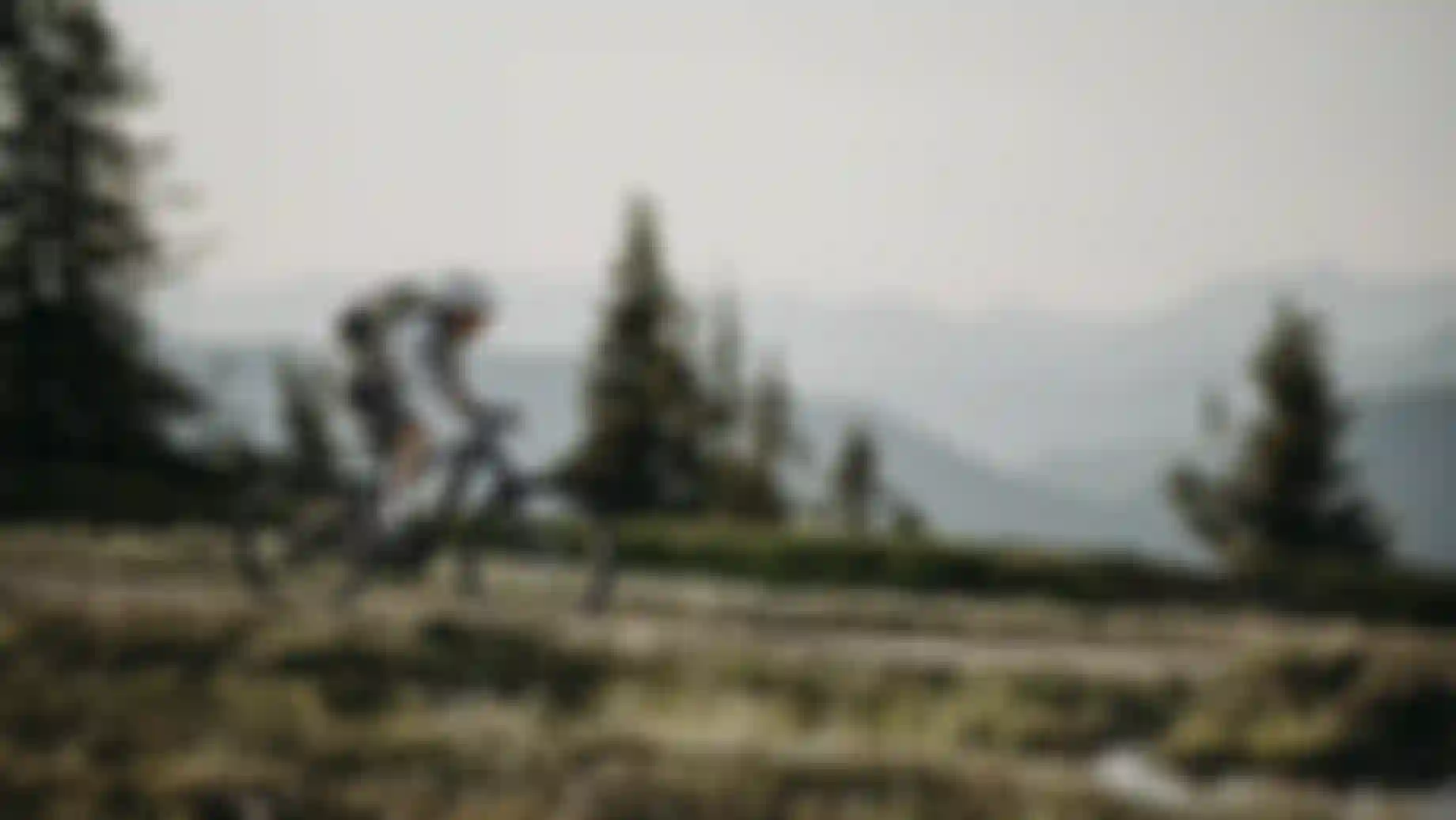 Mountainbiker mit Helm und Rucksack fährt auf einem alpinen Trail durch offene Berglandschaft bei weichem Abendlicht, im Hintergrund verschwommene Bergsilhouetten und Tannen.