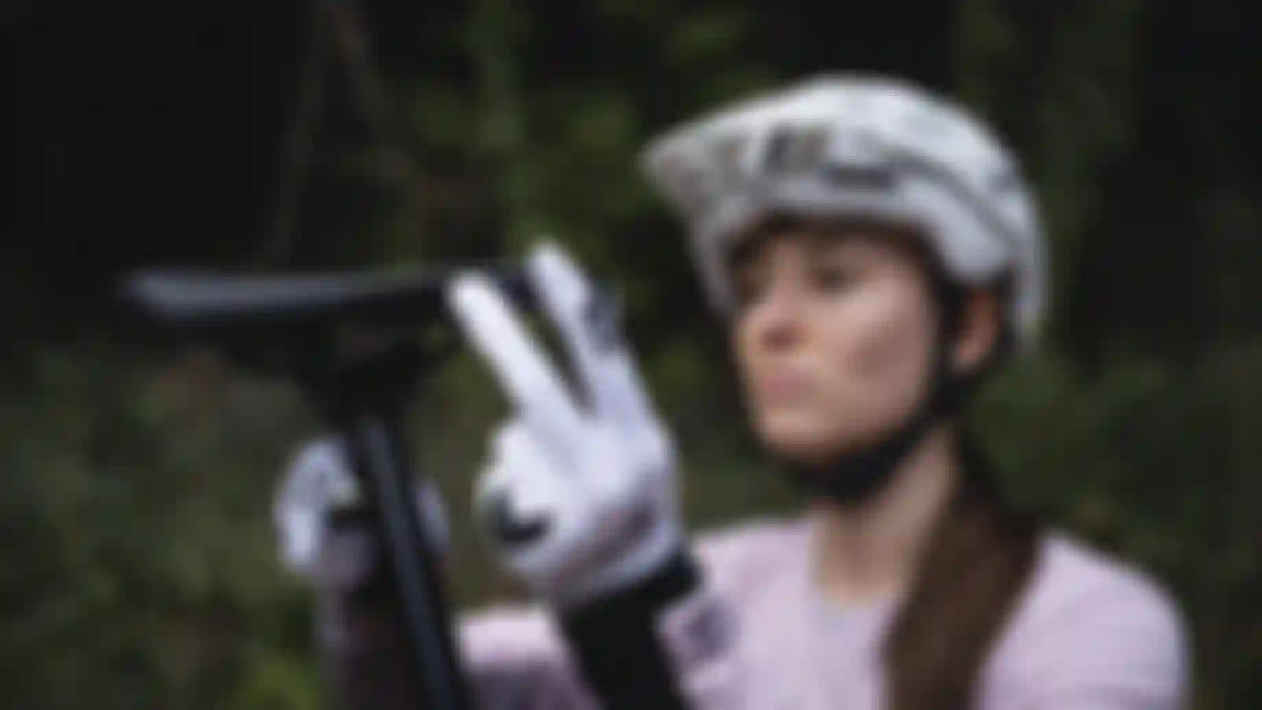 Mountain biker wearing a white helmet and gloves adjusts a bike saddle outdoors, with a blurred forest background.